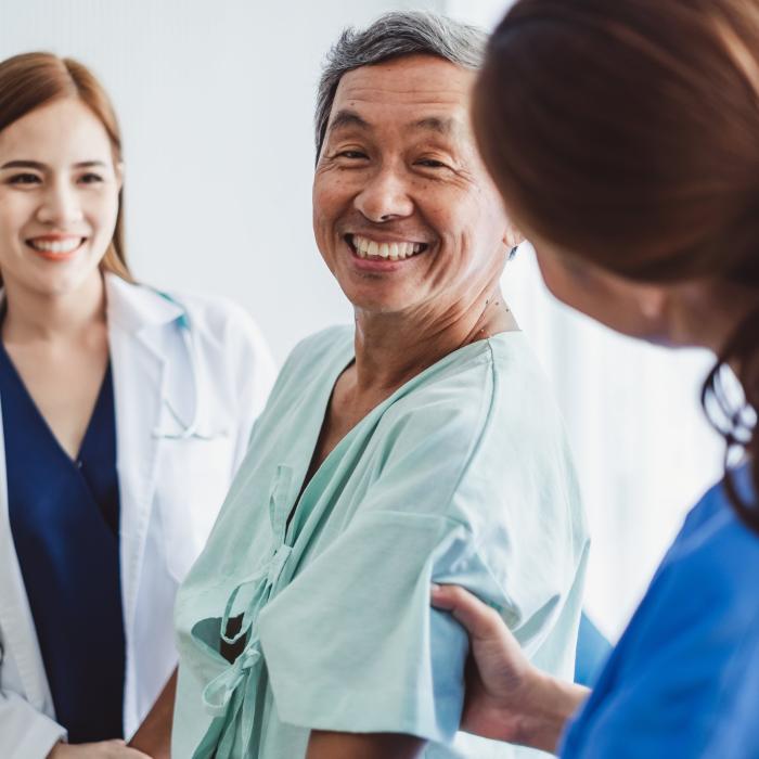A patient is being seen by two nurses at a hospital site. The two nurses are femal with long brown hair, one on each side of the patient who is an older male in a hospital gown. 